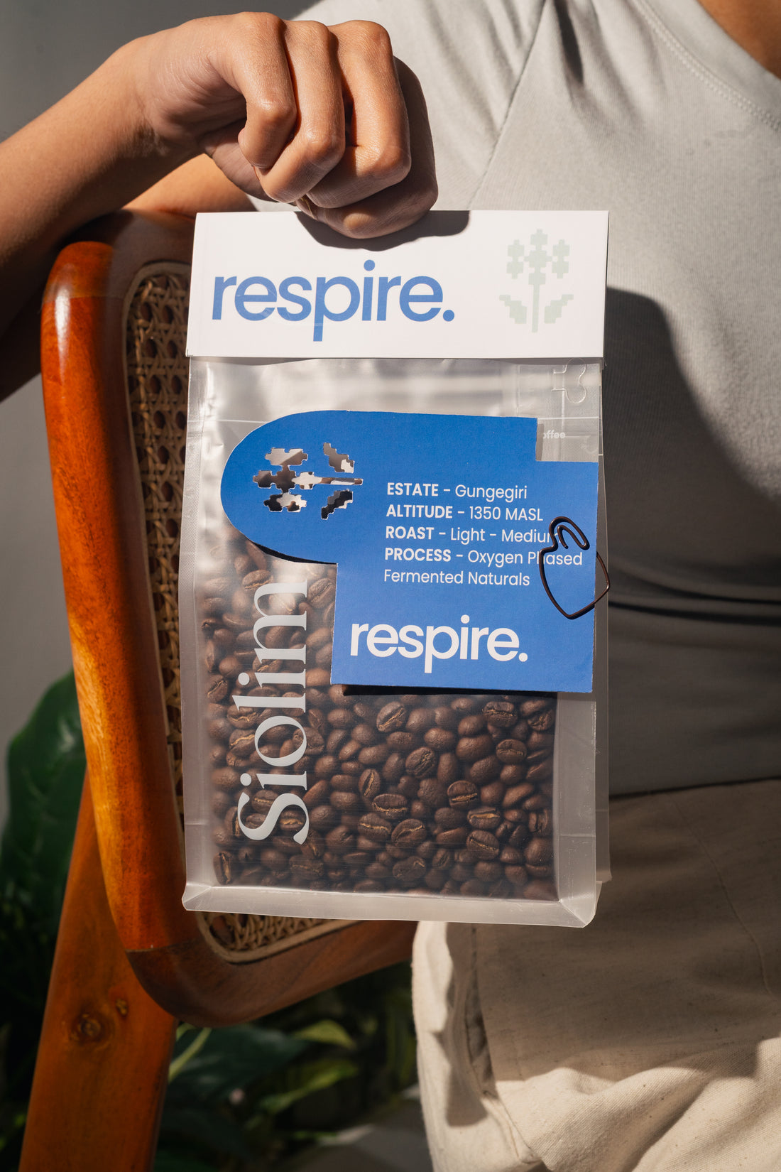 A person holds a clear bag of Respire Solimo Estate coffee against a warm indoor background. The bag is transparent, showing dark roasted coffee beans inside, with white and blue branding labels on the front.